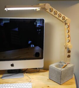 A concrete base with integrated toggle switch anchors the articulating baltic birch ply arm, ending in an LED panel that floods the workspace with light